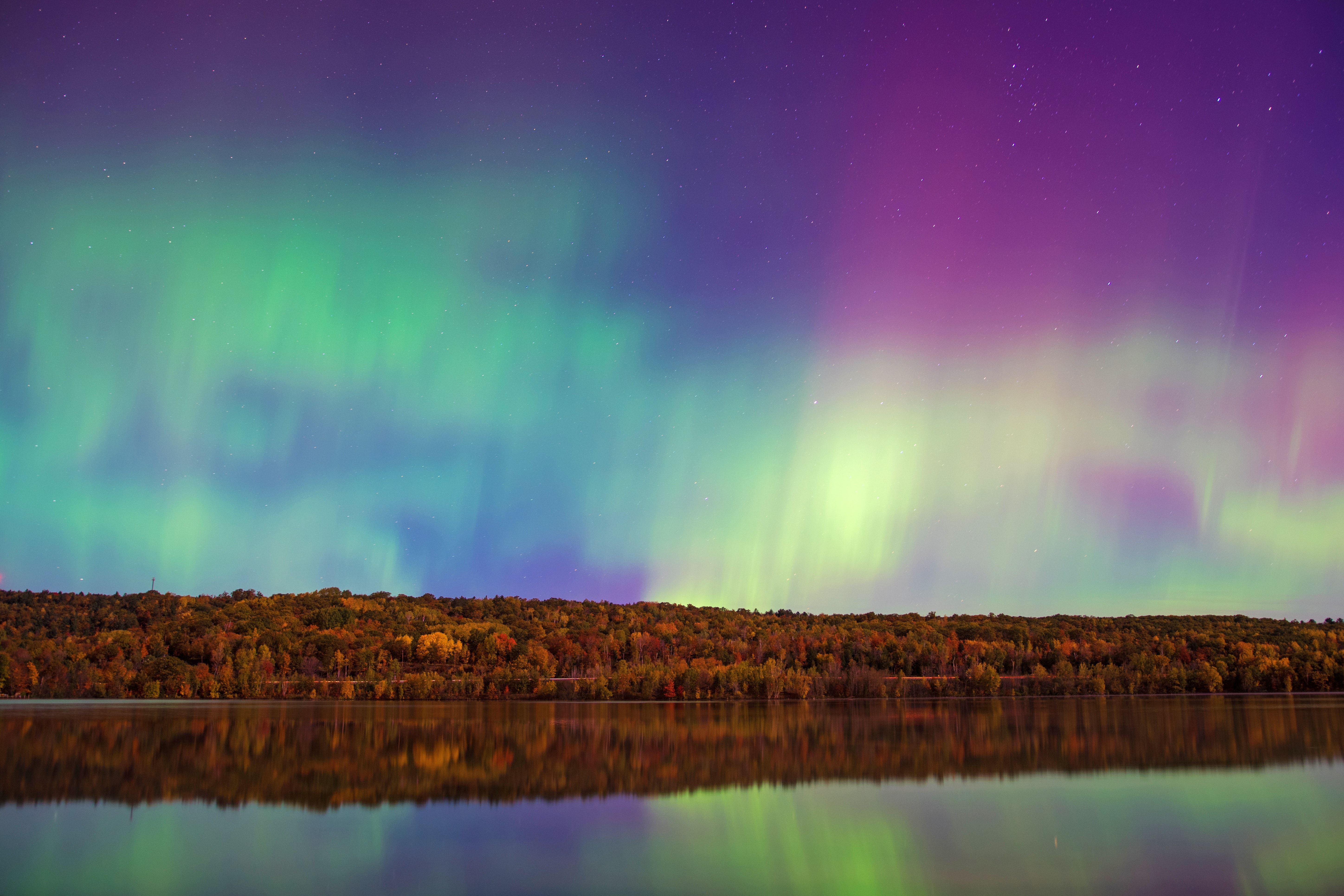 Aurora borealis over trees and a river, made up of green, yellow, pink, and purple. The colors reflect into the river.