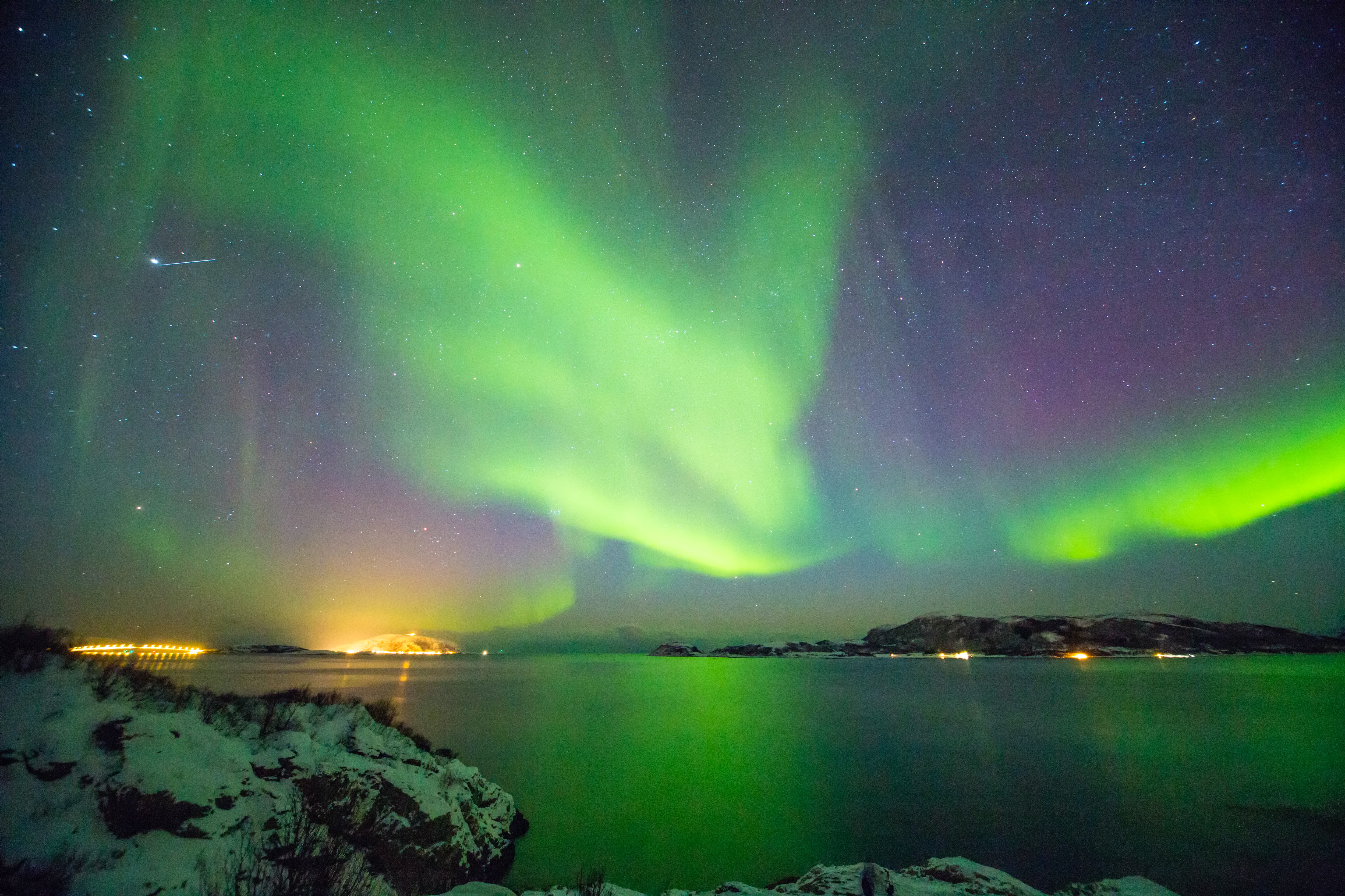 Light green aurora borealis that also has hints of blue and purple over a lake surrounded by snowy mountains.